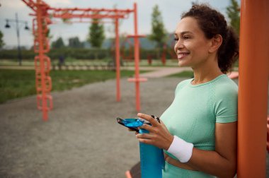 Mutlu muhteşem refah melez Afrikalı Amerikalı orta yaşlı kadın açık hava antrenmanından sonra su içiyor dişlek bir gülümsemeyle barın üzerinde dinleniyor.