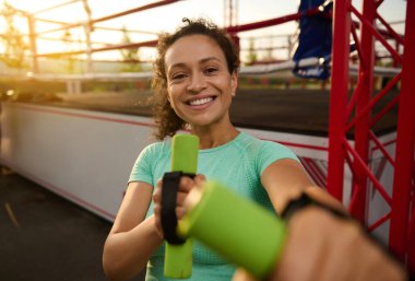 Göz kamaştırıcı Latin Amerikalı çekici genç bir kadının portresi dişlek bir gülümsemeyle, güneşin doğuşunda sabah antrenmanının keyfini çıkarırken, bir boks sahasının arka planında halterlerle poz verirken.