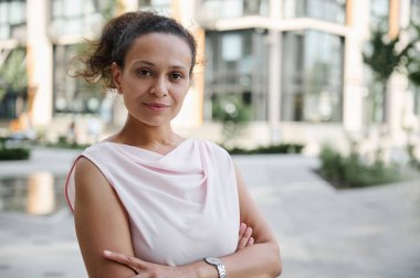Yüksek binaların arka planında çapraz koçlarla kameraya poz veren başarılı, orta yaşlı bir iş kadınının kendine güvenen portresi. Şehir arkaplanı