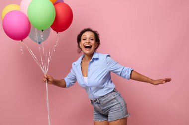 Çekici bir Afro-Amerikan genç kadının bel boyu görüntüsü gülüyor, neşeli, renkli balonlar tutuyor ve kameraya bakıyor, reklam için parlak pembe arka planda poz veriyor.