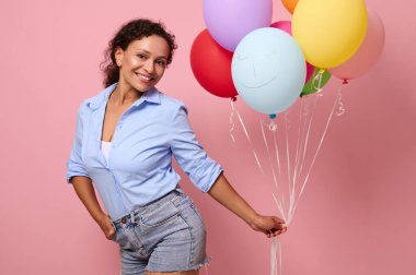 Melez ırk kökenli sevimli genç bir kadın bir sürü renkli balon tutuyor ve pembe arkaplanda mesaj ve reklam için izole edilmiş bir şekilde kameraya bakıyor.