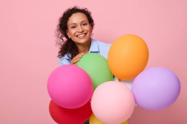 Yakın plan, güzel Afro-Amerikan kıvırcık saçlı, dişlek gülüşlü güzel bir kadın portresi. Renkli balonların arkasında duruyor. Pembe parlak arka planda, fotokopi alanı ile izole edilmiş.