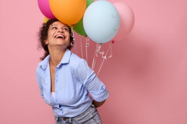 Mavi gömlekli neşeli Afrikalı genç bir kadın dişlek bir gülümseme ile gülüyor ve ellerinde çok renkli hava balonlarına bakıyor. Pembe arkaplan, kopyalama alanı