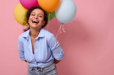 Neşeli ve mutlu duyguları olan genç Latin Amerikalı kadın kameraya bakıp gülüyor, arkasında balonlar tutuyor, pembe renkli arka planda izole edilmiş, reklam için fotokopi alanı olan.