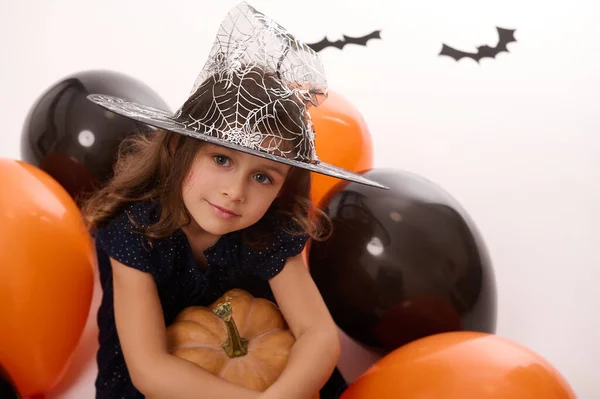 一个戴着魔法帽的漂亮小女孩的画像 穿着深色的女巫狂欢节服装 手里抱着一个南瓜 坐在白色的背景上 戴着五颜六色的黑色气球和蝙蝠 — 图库照片