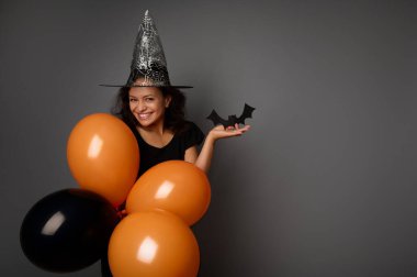 Büyücü şapkalı, siyah giyinmiş, elinde turuncu siyah balonlar ve el yapımı keçeli siyah yarasa tutan güzel, gizemli bir kadın kameraya bakıyor. Gri arka planda Cadılar Bayramı partisi konsepti, kopyalama alanı