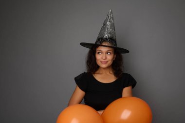Siyah ve cadı şapkalı sevimli gülümseyen Afro-Amerikan kadın turuncu hava balonlarının arasından bakıyor, gri arka planda reklam için bir fotokopi alanına bakıyor. Mutlu Cadılar Bayramı konsepti