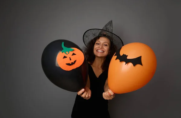 Joyful woman wearing a wizard hat poses with black and orange air balloons with felt-cut bat and pumpkin, smiles toothy smile looking at camera. Halloween concept on grey background, copy space