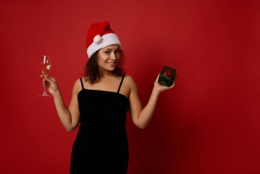 Noel Baba şapkalı ve siyah kadife elbiseli güzel bir kadın köpüklü şarap ve hediye kutusuyla şampanya flütü tutuyor. Kırmızı arka planda gülümseyerek poz veriyor. Noel, Mutlu yıllar