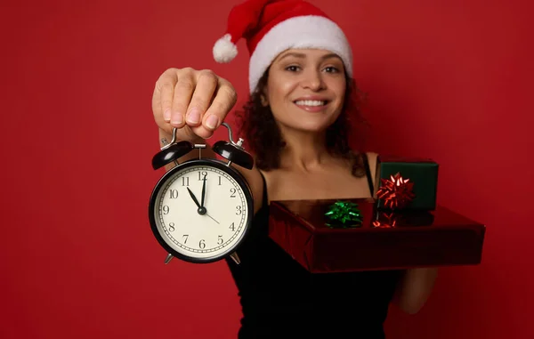 Focus on alarm clock in the hand of blurred cheerful woman in Santa hat, smiling with beautiful toothy smile, holding Christmas gift boxes wrapped in glitter red green gift paper. Copy space for ad