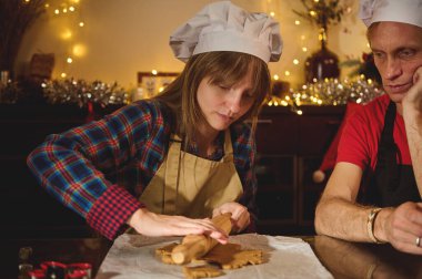 İki yetişkin aşçı şapkası takıyor ve parşömen kaplı bir masada kurabiye kesiyor. Sıcak, şenlikli bir mutfakta bayram hediyesi hazırlıyorlar..