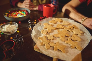 Parşömen üzerinde zencefilli ekmek şekilli kurabiyeler, renkli şekerler ve kurabiye kesicilerle sıcak bir aile sahnesi. Neşe, birliktelik ve evde pişirme aroması..