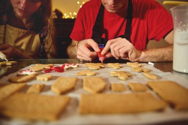 Önlüklü iki kişi kurabiyeleri sıcak, şenlikli bir mutfakta kremayla süslüyor. Aile dostu bir bayram pastası için parşömen üzerine yazılmış kurabiyeler..