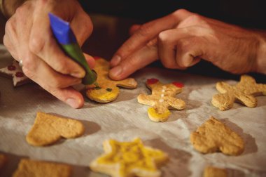 Çeşitli şekerli kurabiyeleri parşömen üzerine renkli kremalarla süsleyerek tatil pişirme sırasında sıcak, şenlikli bir aile anını yakalayan ellerin yakın çekimi..