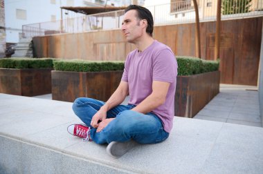 Rahat bir adam, şehir avlusunda beton bir çıkıntıda bağdaş kurmuş, mor bir gömlek ve kot pantolon giyiyor. Paslı ekiciler ve temiz çizgiler modern ve düşünceli bir hava yaratıyor..