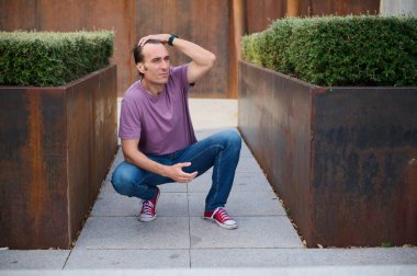 Mor tişörtlü ve kot pantolonlu bir adam paslı metal ekicilerin arasına çömelmiş yeşil çalılar, açık hava, gösterişli moda, sokak tarzı, ve rahat ruh hali.