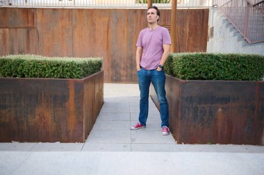 Pembe tişörtlü ve kot pantolonlu bir adam, elleri paslı saksıların arasında duruyor ve açık hava ortamında çitleri buduyor, gündelik güveni ve sokak tarzını aktarıyor..