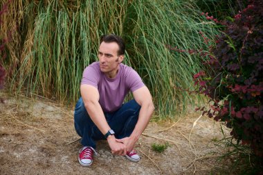 Mor t-shirt ve kot giyen bir adam uzun otların ve çalıların arasında kuru zemine çömelir, sakin ve açık hava sporu anı yaşar ve odaklanır..