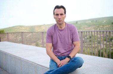 Mor tişörtlü bir adam beton bir çıkıntıda oturur, sakin ve düşünceli bir ifadeyle mesafeye bakar. Manzaralı tepeler ve korkuluklar sakin bir arka plan sağlar..