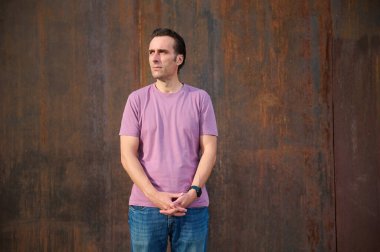 Pembe tişörtlü rahat bir erkek model paslı, desenli bir duvarın önünde duruyor, moda, yaşam tarzı ve editoryal stok tasvirleri için sakin bir güven ve kentsel tarzı iletiyor..