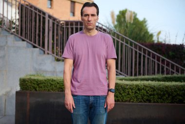 Pembe tişörtlü ve kot pantolonlu bir adam merdiven ve manikürlü çitlerin yanında dikiliyor. Yaşam tarzı, moda ya da sağlık konularına uygun sakin, sıradan bir portre..