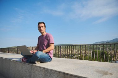 Bir adam beton bir çıkıntıda oturuyor, açık mavi gökyüzü ve uzak tepeleri olan bir dizüstü bilgisayarda yazıyor. Uzaktan çalışma, seyahat ve açık hava verimliliği için ideal.
