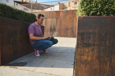Mor gömlekli ve kot pantolonlu bir adam paslı duvarların yanına çömelmiş, şehir avlusunda dizüstü bilgisayar kullanıyor, uzaktan çalışma, odaklanma ve günlük verimliliği aktarıyor..