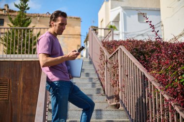 Sıradan bir profesyonel, elinde dizüstü bilgisayarla merdivenlerde durup akıllı telefonunu kontrol eder. Parlak şehir manzarası uzak çalışma, bağlantı ve modern, üretken bir yaşam tarzı sunar..