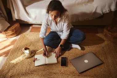 Bir kadın evdeki dokuma halıya bağdaş kurup dizüstü bilgisayarı ve telefonu yakınlarda dinlenirken deftere bir şeyler yazıyor. Kahve fincanı rahat ve verimli bir dokunuş ekler..