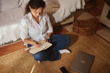 Bir kadın, dizüstü bilgisayarı ve akıllı telefonu yakınlarda dinlenirken örülmüş bir halıya bağdaş kurup bir deftere yazı yazar..