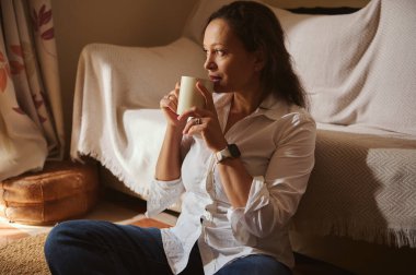Rahat bir kadın yerde, rahat bir kanepenin yanında oturuyor, bardaktan yudumluyor. Evde sakin ve rahat bir anın tadını çıkarırken güneş ışığı odayı dolduruyor..