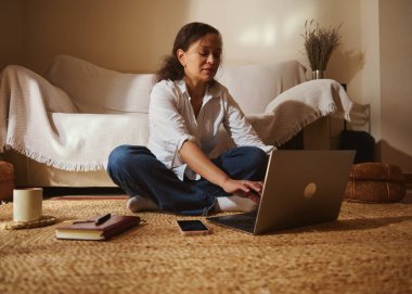 Bir kadın, sıcak, rahat bir oturma odasında dizüstü bilgisayarının üstünde bağlı bir şekilde oturuyor. Yumuşak ışık ve sakin ruh hali ev işlerini ve odaklanmayı iletir..