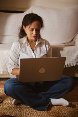Bir kadın evde dizüstü bilgisayarla halının üzerinde bağdaş kurarak oturuyor. Rahat, odaklanmış ev hayatını rahat ve rahat bir ortamda işle harmanlayan bir an..