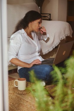 Beyaz tişörtlü bir kadın evde dizüstü bilgisayarda çalışıyor, kahve fincanıyla halıya oturmuş, uzaktan kumandalı rahat bir iş sahnesi yaratıyor..