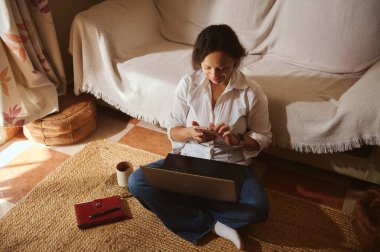 Bir kadın güneşli bir oturma odasında dizüstü bilgisayar ve akıllı telefon kullanarak halının üzerinde bağdaş kurarak oturuyor. Sahne, uzaktan çalışma, odaklanma ve rahat bir çalışma alanı taşır.