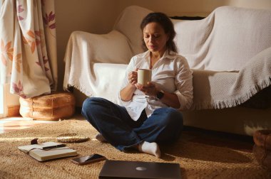 Bir kadın, elinde kupayla güneşli bir oturma odasında, rahat bir halının üzerinde bağdaş kurarak oturuyor. Laptop, dizüstü bilgisayar ve telefon yakınlarda, sakin bir ev ofisi anı..