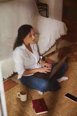 Oturma odasında rahat bir yerde oturan bir kadın dizüstü bilgisayarında bir defter, kupa ve telefonun yanında yazı yazıyor. Uzaktan çalışma, ev ofisi, çalışma ve verimlilik görselleri için ideal.