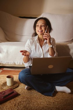 Neşeli bir kadın evdeki dokuma halıda dizüstü bilgisayar kullanarak ve akıllı telefonla konuşarak bacak bacak bacak üstüne atarak oturuyor. Sıcak gün ışığı rahat, modern bir ofis ortamı yaratır..