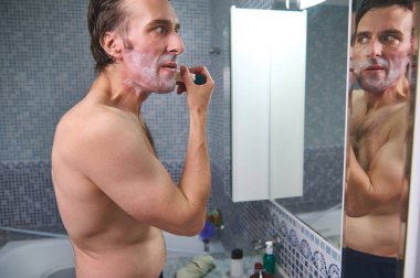 Tişörtsüz bir adam banyo lavabosunda tıraş olur, aynaya bakarken köpük sürer. İçinde bakım ürünleri olan fayanslı bir banyo gündelik, kişisel bakım anlarını yakalar..