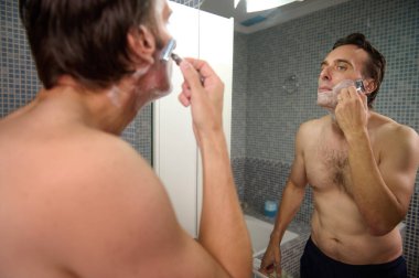 Üstsüz bir adam traş kremini fayanslı bir banyoda jiletle sürüyor, aynada sakin bir bakım sırasında ve kişisel bakım anında görülüyor..