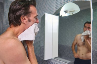 Üstsüz bir adam yüzünü fayanslı bir banyoda havluyla kurutuyor. Yandan aynadaki yansımasıyla görülüyor. Sahne rutin tımar ve sakin sabah anlarını aktarıyor..