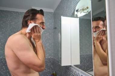 Üstsüz bir adam fayanslı bir banyoda yüzünü havluyla siliyor, aynada yansıması görünüyor. Samimi bir bakım ve bakım anı..