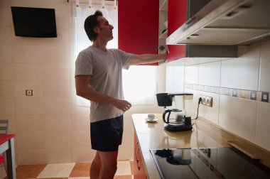 Gri tişörtlü ve şortlu bir adam bir kahve makinesinin üstündeki kırmızı mutfak dolabını açar ve çağdaş bir ev mutfağında içki hazırlar..