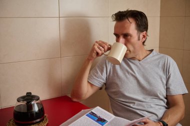 Gri tişörtlü rahat bir adam fayanslı bir mutfakta kırmızı masada dergi okurken kahvenin tadını çıkarıyor. Çaydanlık her sabah daha rahat bir hava katar..