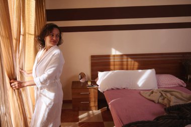 Beyaz bornozlu rahat bir kadın güneşli bir pencerenin yanında durur, çizgili perdeleri çekerek rahat bir yatak odasını, yastıkları ve yumuşak bir battaniyeyi ortaya çıkarır..