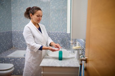 Beyaz bornozlu bir kadın ellerini mavi mozaik fayanslarla çevrili banyo lavabosunda yıkıyor ve sakin, temiz bir spa havası yaratıyor..