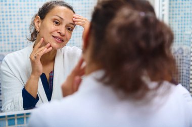 Bir kadın banyo aynasına cilt kremi sürerken yansımasına gülümsüyor. Bu samimi an kişisel bakım, günlük rutin ve sağlıklı güzellik uygulamalarını yakalar..