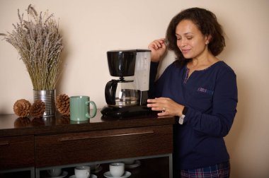 Bir kadın, lavanta, kozalak ve yeşil kupalarla çevrili, tahta bir tezgahın üzerinde sade bir kahve makinesinin yanında duruyor. Sıcak ve sakin bir ev sahnesi..