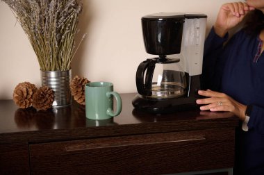 Sıcak bir mutfak düzeneği. İçinde damlayan kahve makinesi, yeşil kupa, kozalak ve koyu ahşap tezgahta kurutulmuş lavanta var. Biracının yanındaki eller sakin bir sabah rutini olduğunu gösteriyor..