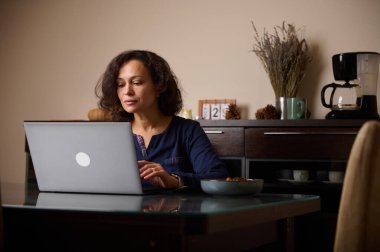 Ev mutfak masasında dizüstü bilgisayara odaklanmış bir kadın oturuyor. Sahnede bir kahve makinesi, kupa ve kâseler, sakin ve üretken bir çalışma anı sunuluyor..